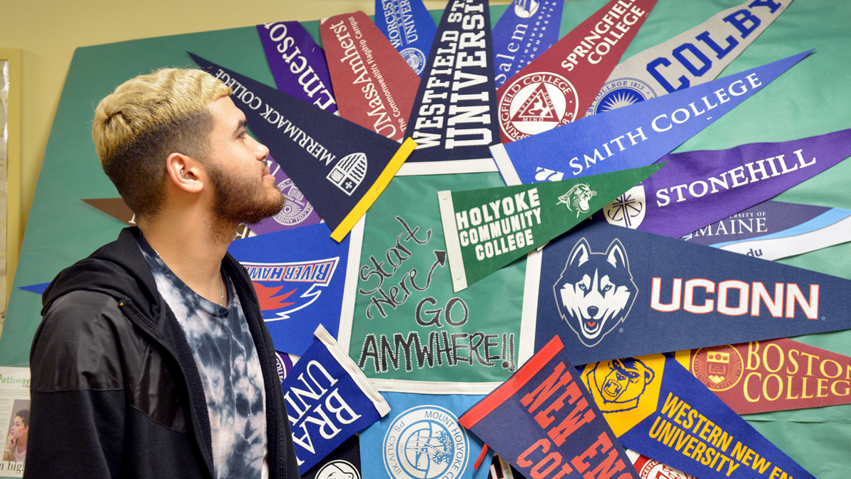 A male student looks at a bulletin board display made up of pennant flags from a variety of four-year schools and universities